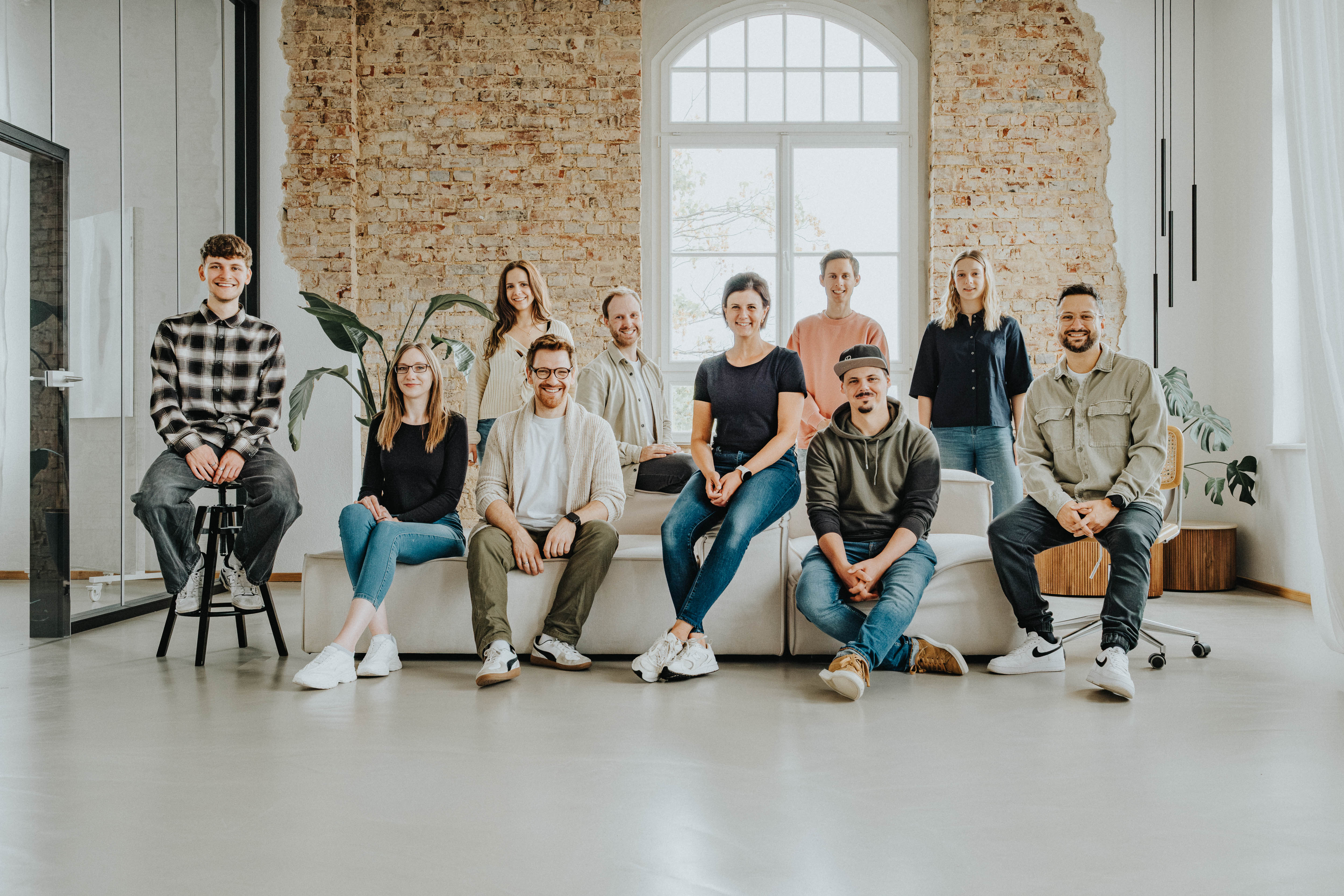 Zehn Personen sitzen und stehen gemeinsam in einem hellen Büro mit Backsteinwand und großem Fenster; sie posieren als Teamfoto auf und vor einem Sofa.