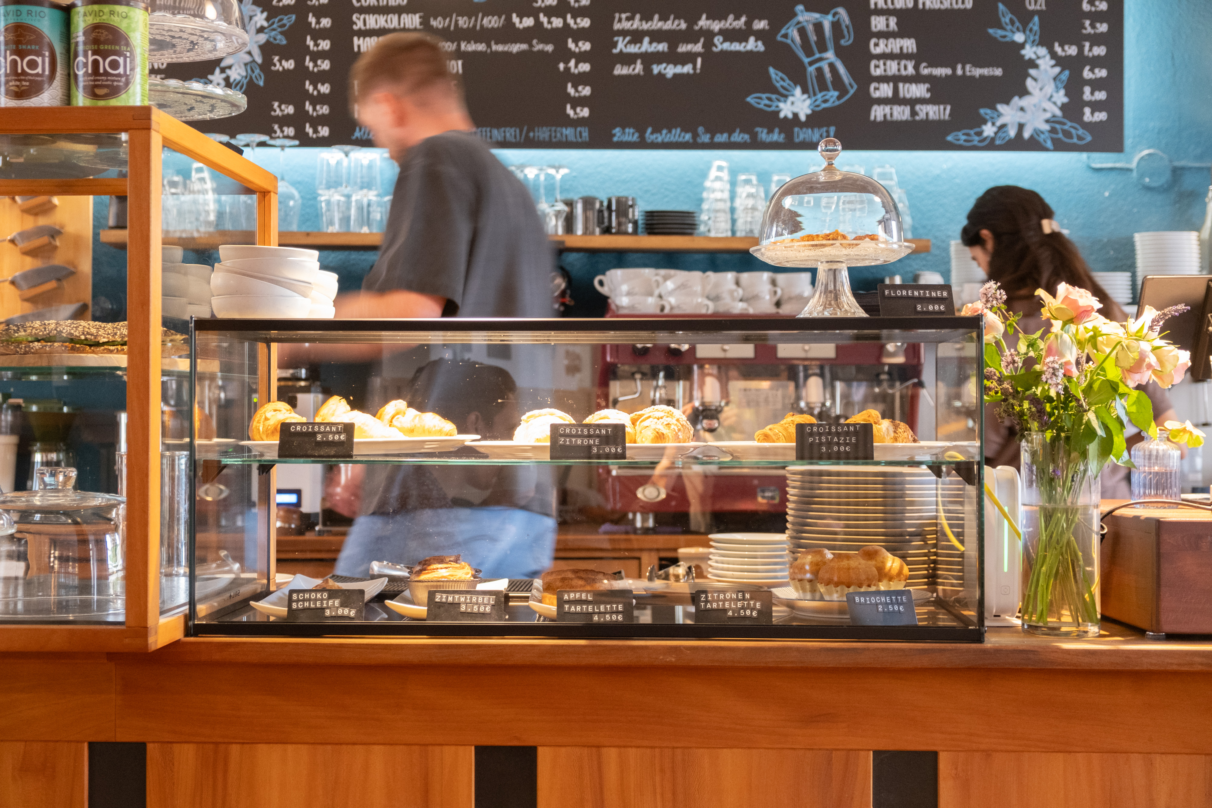 Theke eines Cafés mit Glasvitrine voller Croissants und Tartes, Barista im Hintergrund vor einer beschrifteten Kreidetafel.