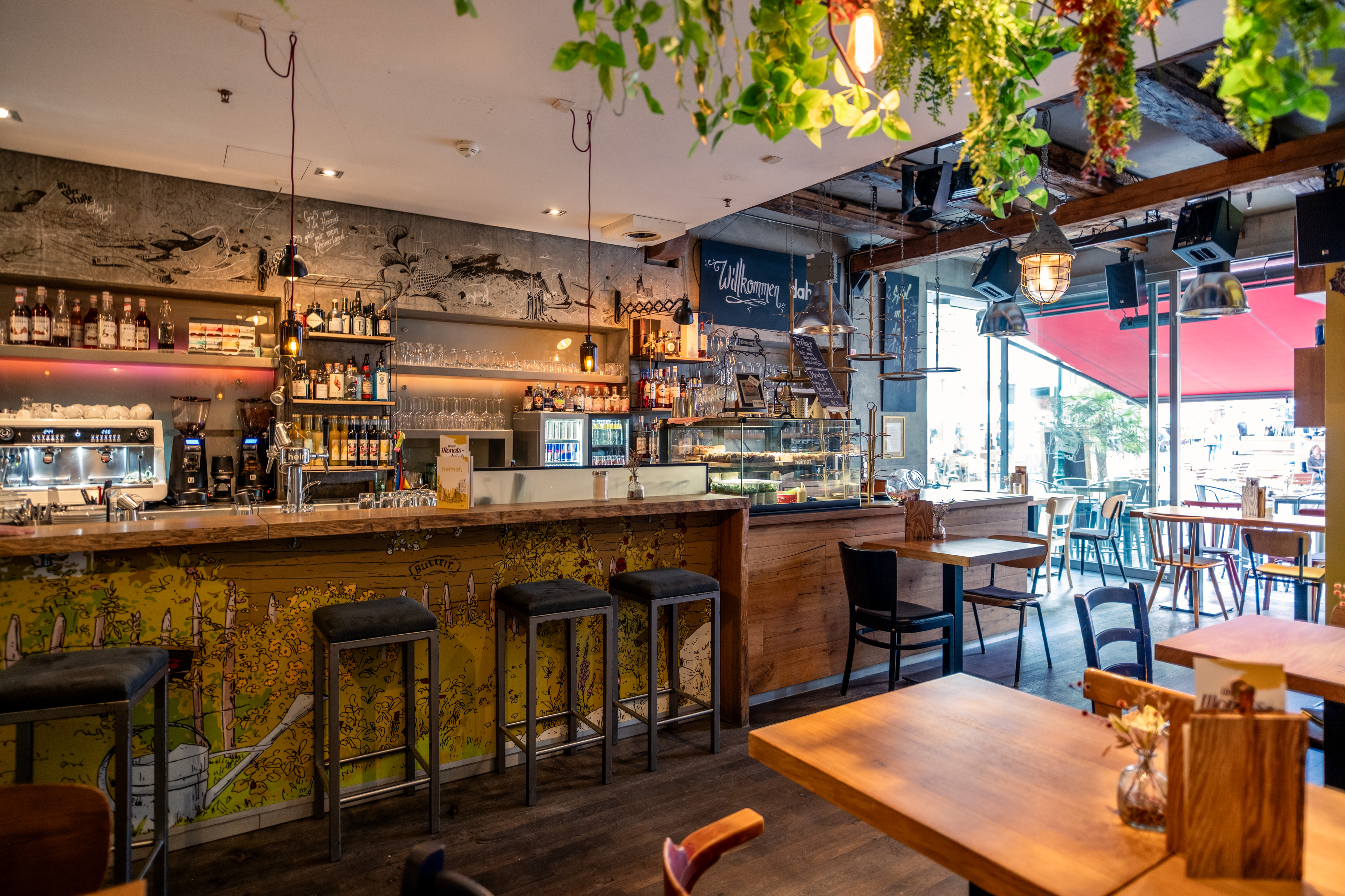 Gemütliches Café mit Holztischen, Barbereich mit Espressomaschine und Spirituosenregal, hängenden Pflanzen und großen Fenstern zum Außenbereich.