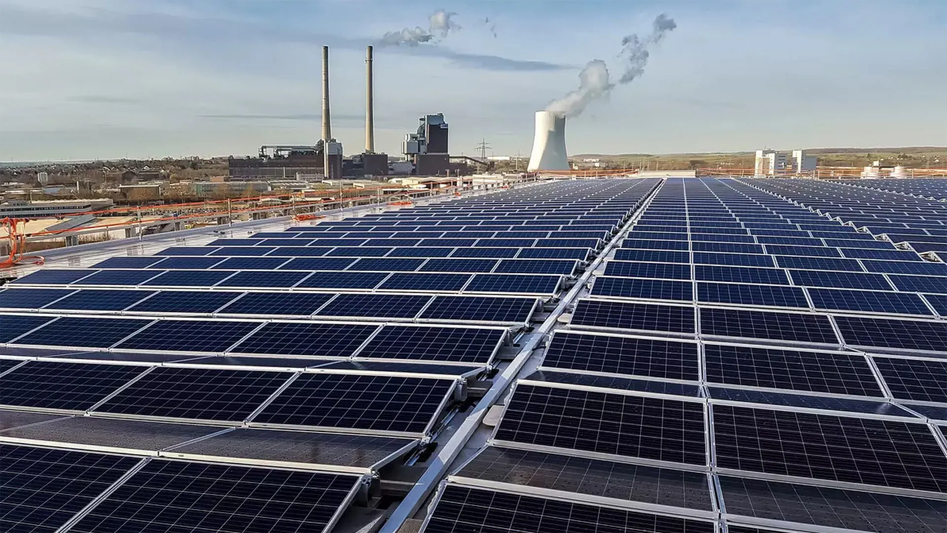 Rows of solar panels installed on a rooftop with an industrial power plant and smokestacks emitting steam in the background.