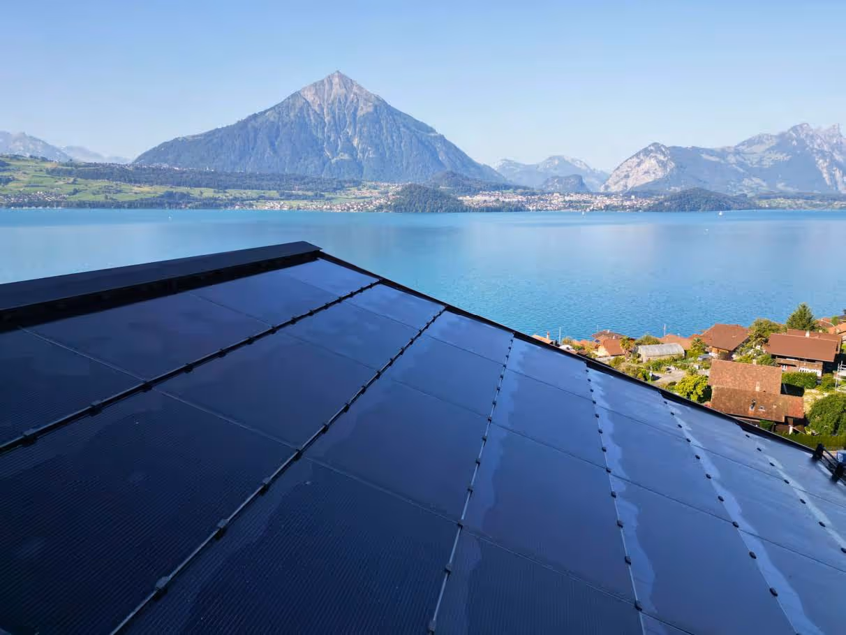 Landschaftsfoto aus Luftperspektive, zeigt vier Mehrfamilienhäuser mit Solaranlagen auf den Flachdächern, im Hintergrund Thunersee und Bergpanorama.