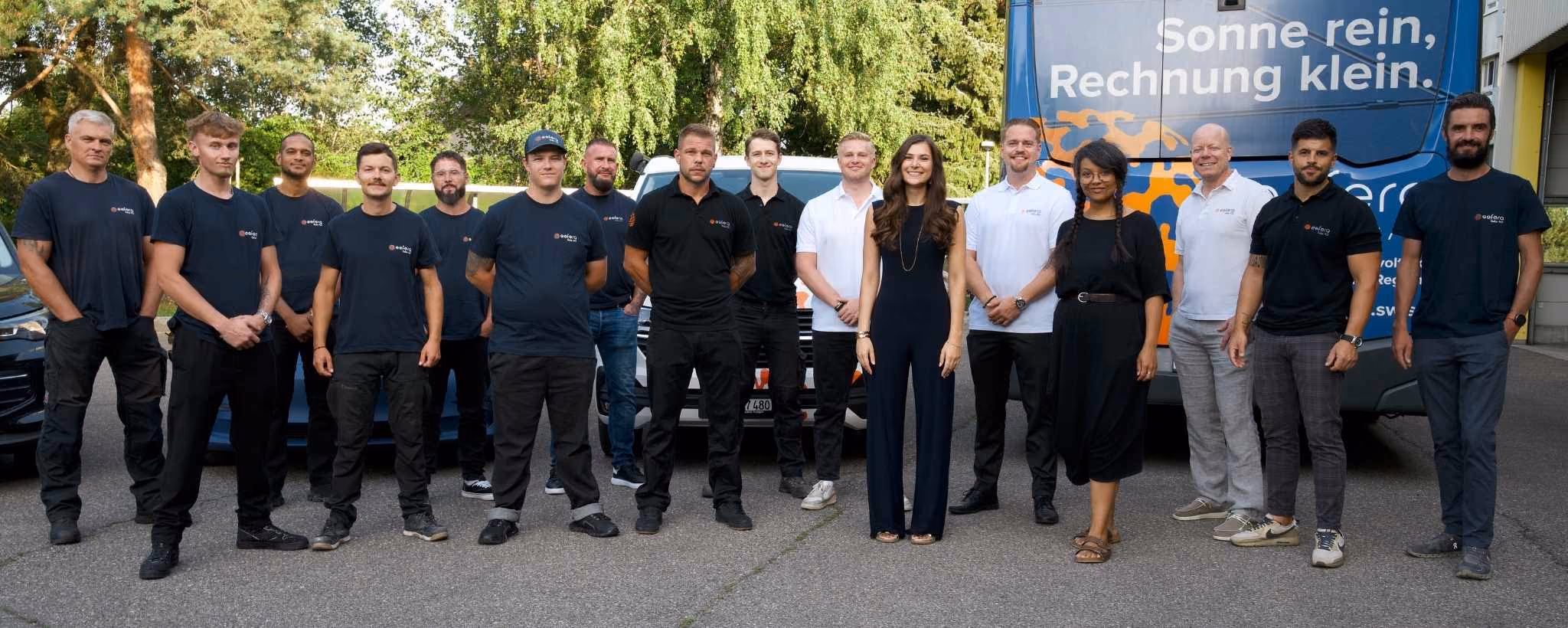 Group of 16 diverse people of the esfera team in casual work attire standing outdoors between a custom Volkswagen car and a blue STI bus with 'Sonne rein, Rechnung klein.' text.
