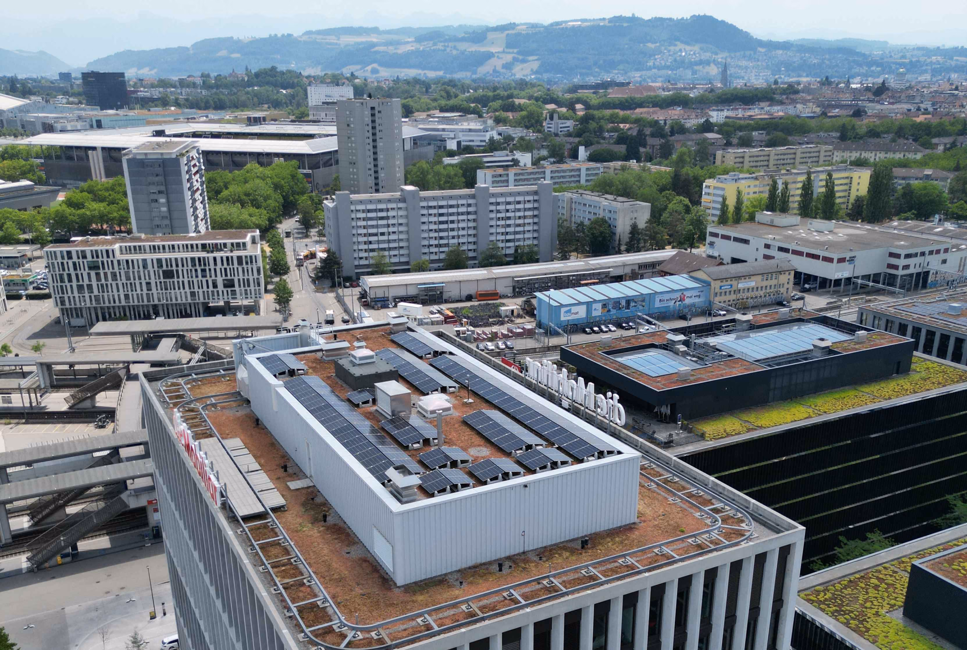 Landschaftsfoto aus Luftperspektive, zeigt vier Mehrfamilienhäuser mit Solaranlagen auf den Flachdächern, im Hintergrund Thunersee und Bergpanorama.
