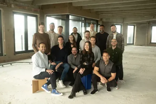 Group of sixteen casually dressed people posing and smiling in an unfinished office space with concrete floors and large windows.