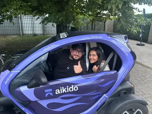 Two people sitting inside a small purple electric car branded with 'aikido', both showing thumbs up.