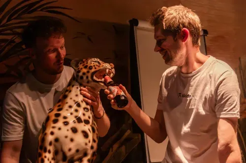 Two men, one holding a large leopard statue while the other feeds it a bottled drink, both smiling indoors.