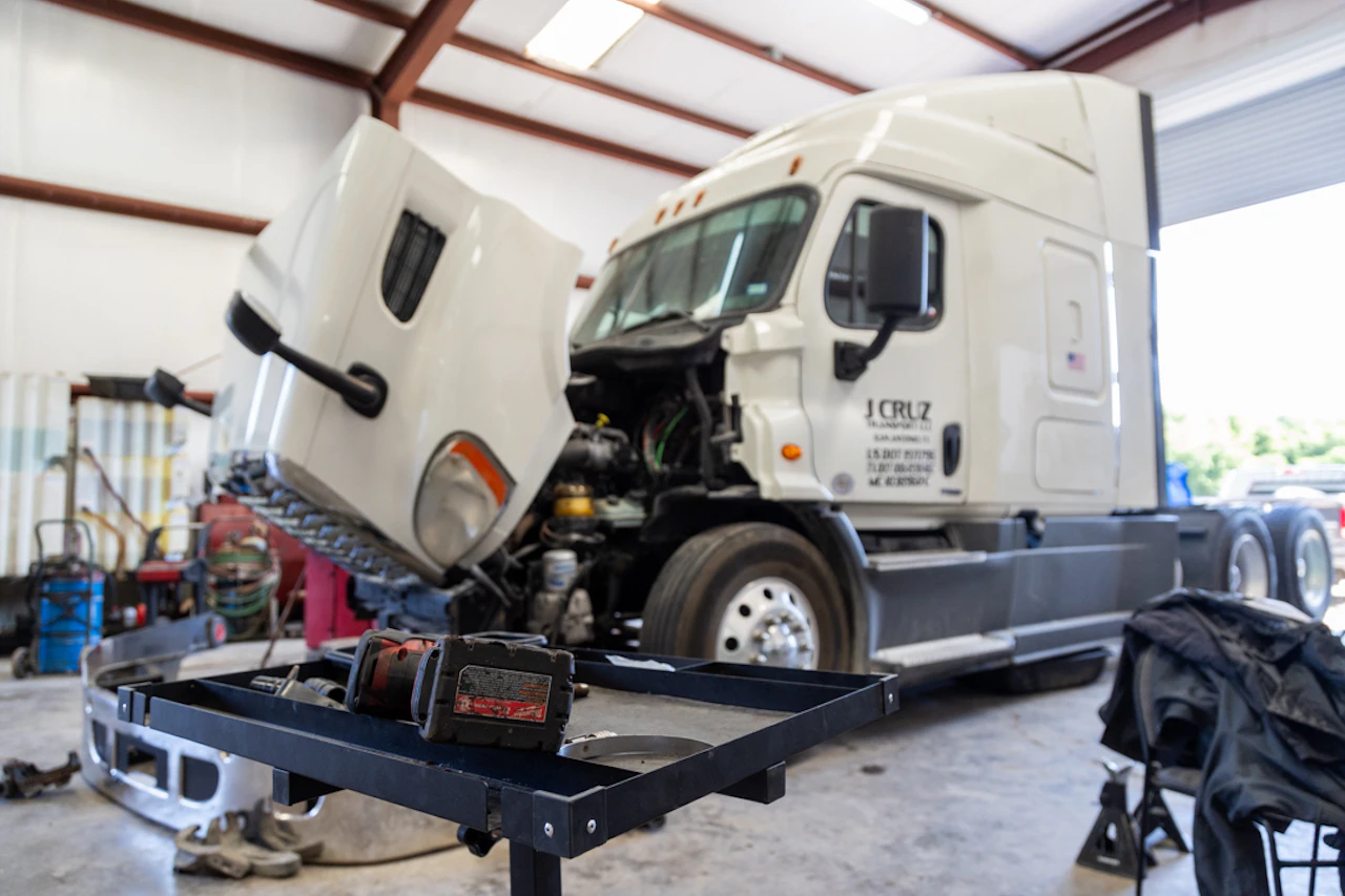 A white semi-truck with its hood raised is undergoing maintenance inside a well-lit garage, with tools and parts scattered around the floor. In the foreground, a black workbench holds a cordless power tool, emphasizing the active repair environment.