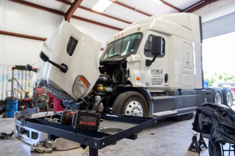 A white semi-truck with its hood raised is undergoing maintenance inside a well-lit garage, with tools and parts scattered around the floor. In the foreground, a black workbench holds a cordless power tool, emphasizing the active repair environment.