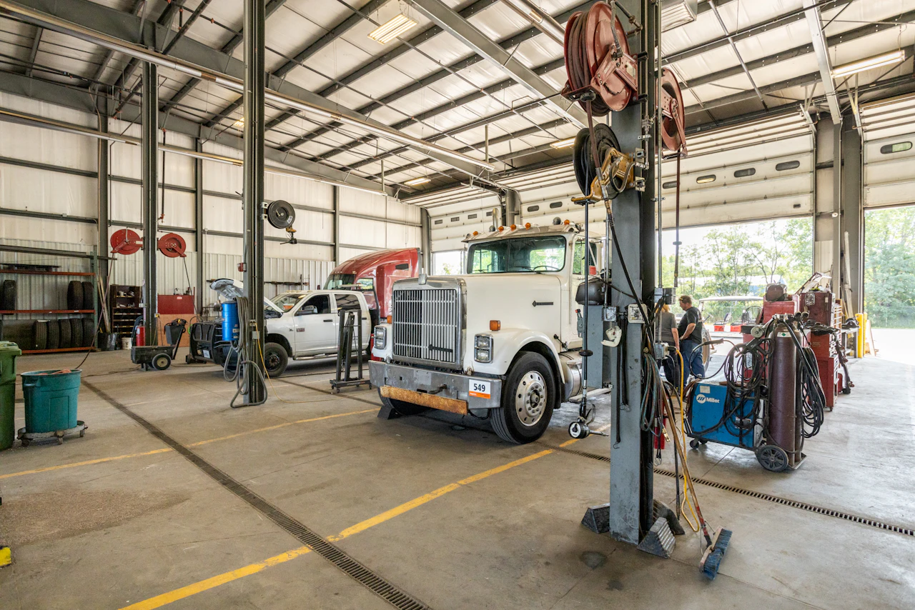 Inside a spacious and well-lit vehicle maintenance garage, a white semi-truck, a white pickup truck, and a red semi-truck cab are positioned in service bays surrounded by tools, equipment, and hose reels. Two people stand near the open garage door, talking beside welding tanks and machinery.