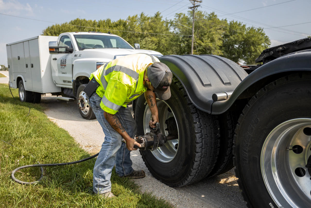 A roadside mechanic in a neon yellow safety shirt and baseball cap uses a pneumatic impact wrench to service the rear tire of a large truck. A white B&B service vehicle is parked nearby on a rural road bordered by grass and trees under a clear sky.