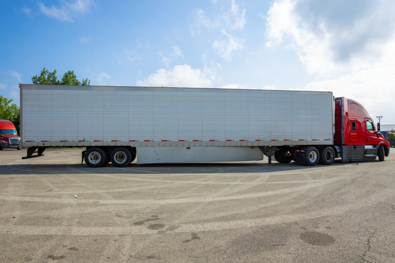 A red semi-truck with a long white trailer is parked on a wide asphalt lot under a bright blue sky. Another red truck is partially visible in the background near trees on the left.