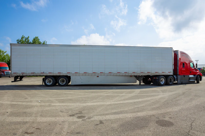 A red semi-truck with a long white trailer is parked on a wide asphalt lot under a bright blue sky. Another red truck is partially visible in the background near trees on the left.