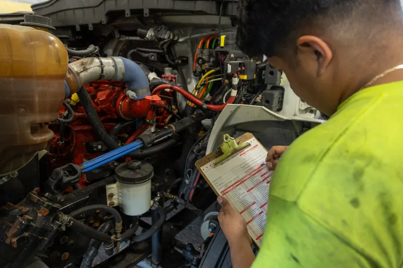 Technician diagnosing diesel truck problems while inspecting engine bay and checking a clipboard with maintenance checklist.