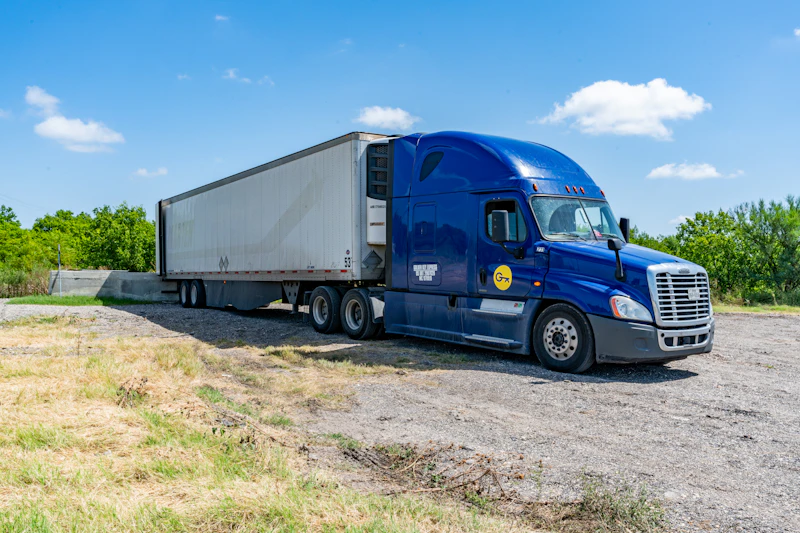 Fleet service showing blue semi truck with refrigerated trailer parked on gravel lot under clear sky.