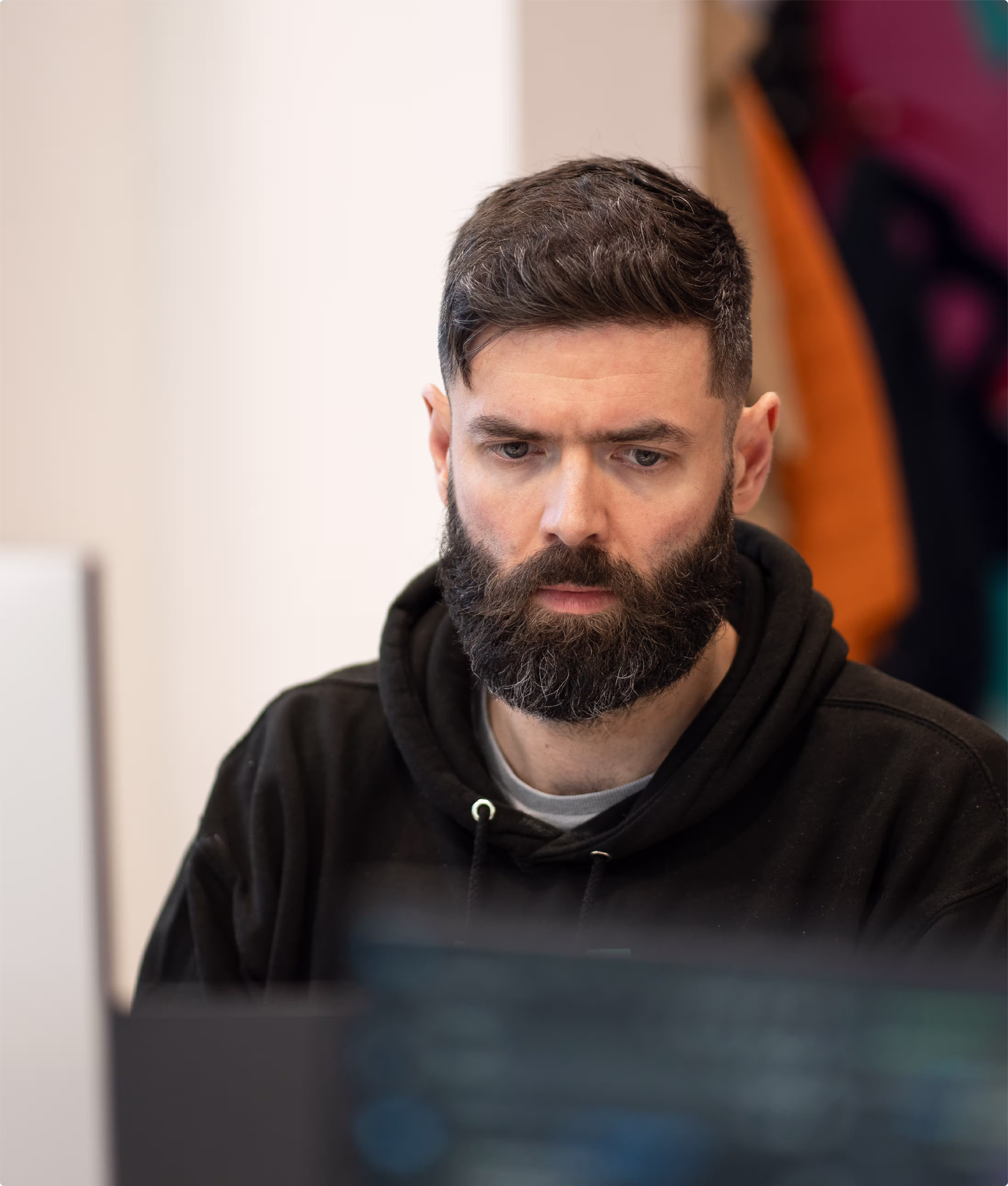 Bearded man in a black hoodie focused on a computer screen with blurred code in the foreground.