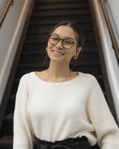 Girl with glasses and white sweater Infront of the bottom steps of a staircase