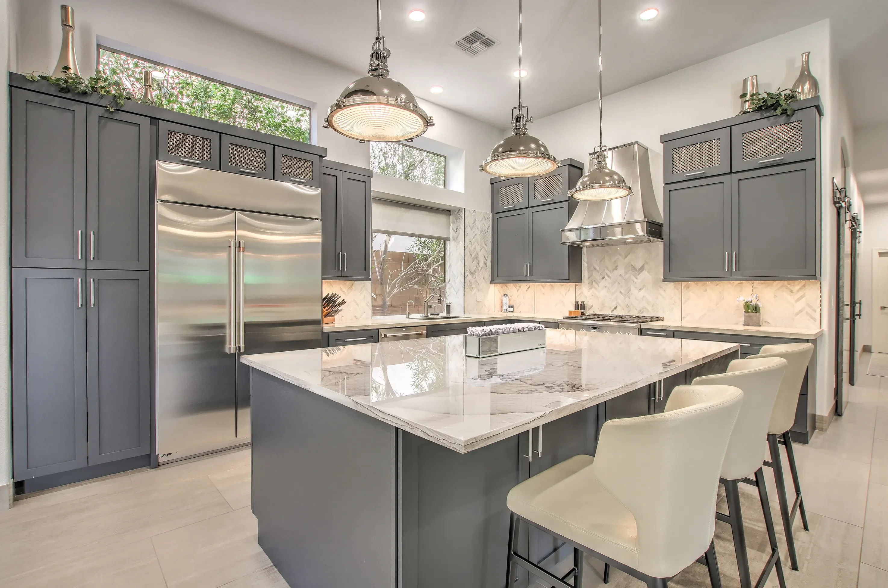 Modern kitchen with gray cabinetry, large marble island, stainless steel appliances, and three cream-colored bar stools.