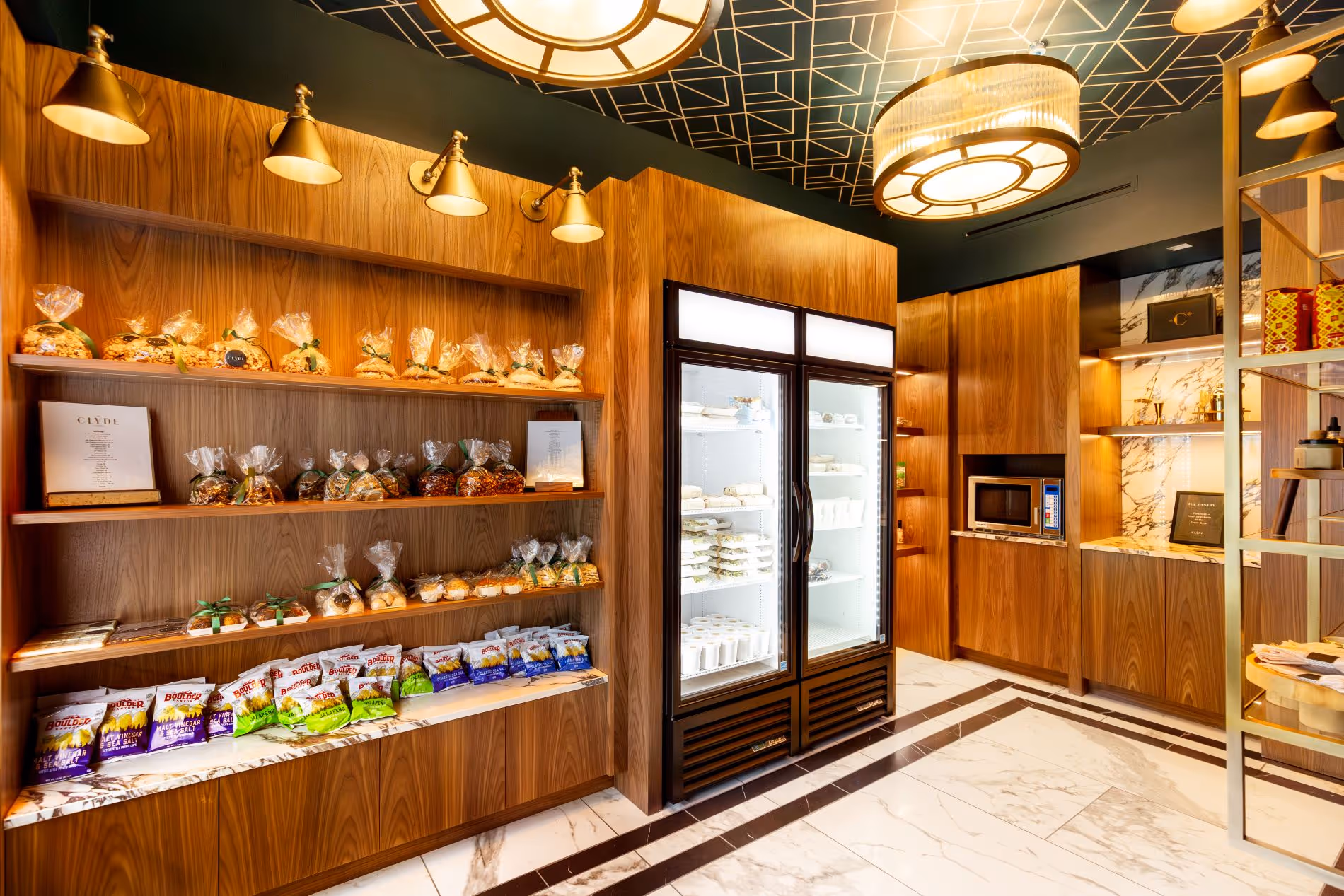 The Clyde Pantry with marble floors green geometric patterned ceiling, gold lights and wooden walls, along with a cooler filled with grab and go food items and a shelf with various snack items for purchase
