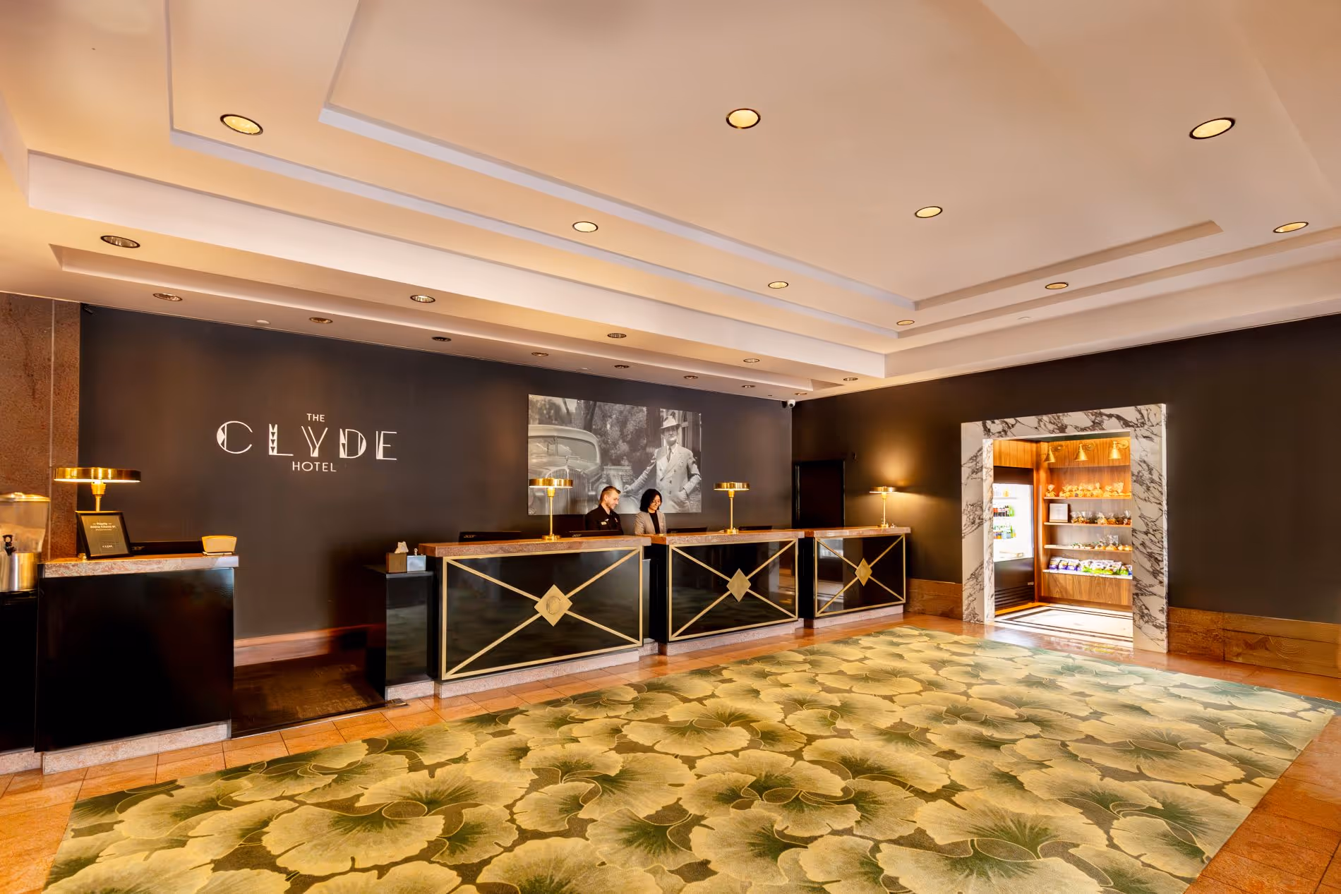 Front desk of The Clyde Hotel with green gingko patterned carpet black wall behind the front desk with a picture of Clyde Tingley standing next to a car and various gold accents on the desk and lamps on the desk