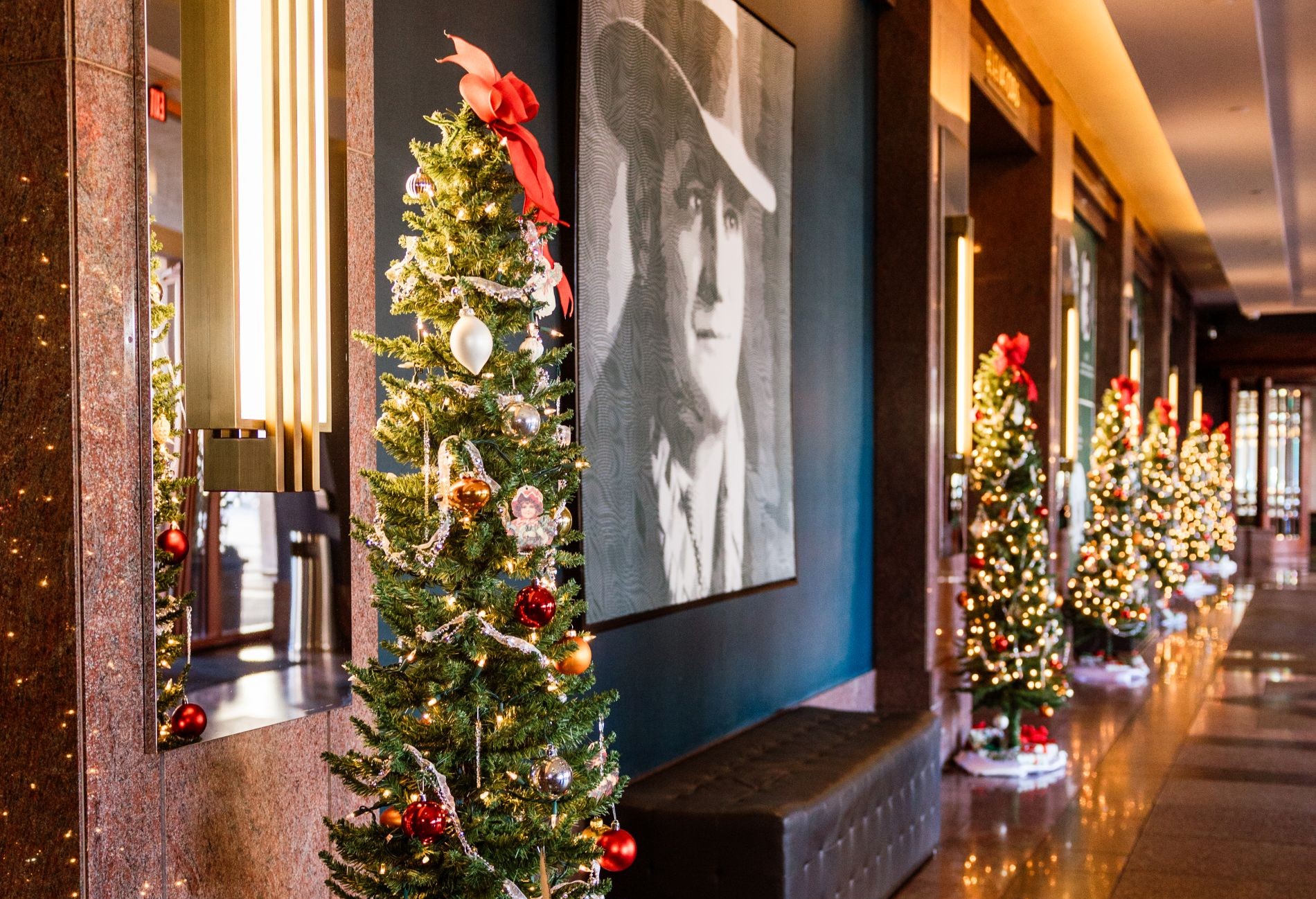 Several decorated Christmas trees down a hall at Clyde with a image of Clyde on the wall