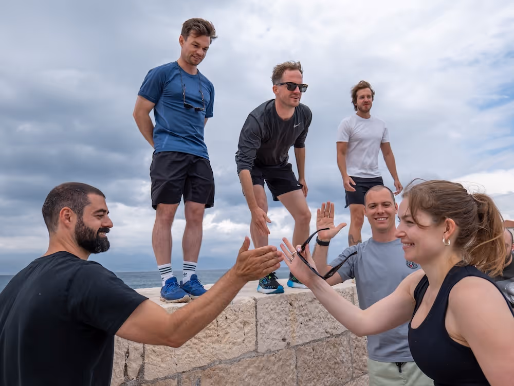 Six young adults outdoors on a stone wall against a cloudy sky, two in the foreground giving a high-five and others standing or crouching behind them, all casually dressed in athletic wear.
