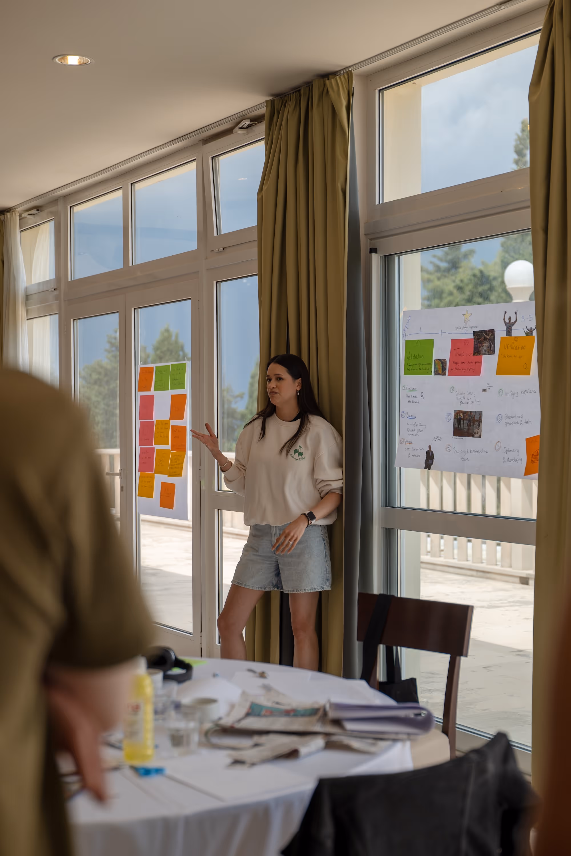 Woman standing near glass doors presenting ideas on sticky notes and posters during a meeting.
