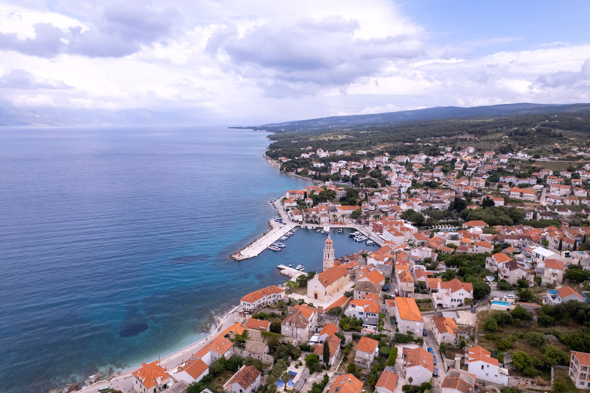 Aerial view of a coastal town with terracotta rooftops, a harbor with boats, and calm blue sea under a cloudy sky.