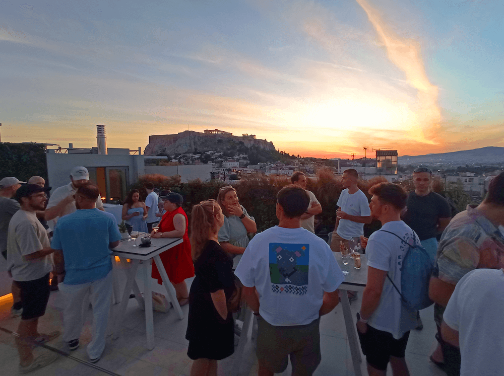 A group of people at a rooftop party in Athens during sunset, with the Acropolis visible on a hill in the background.