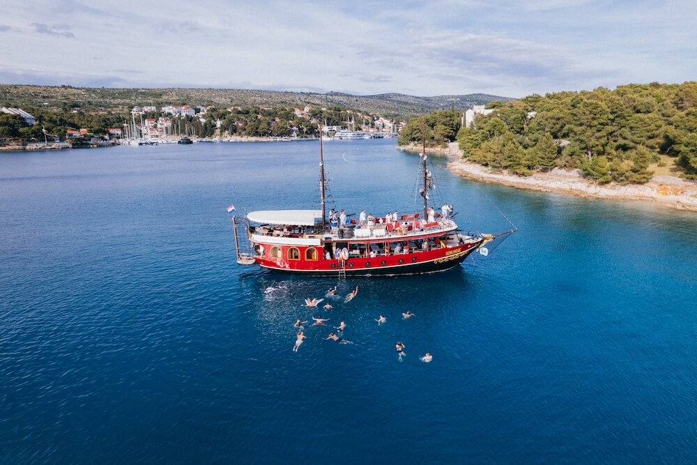A traditional red excursion boat anchored in a blue bay with people swimming in the water nearby.