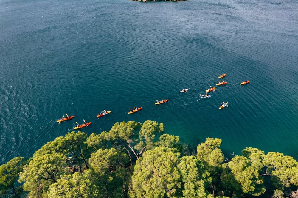An aerial view of a group of people paddling colorful kayaks in a line across deep blue water near a tree-lined shore.