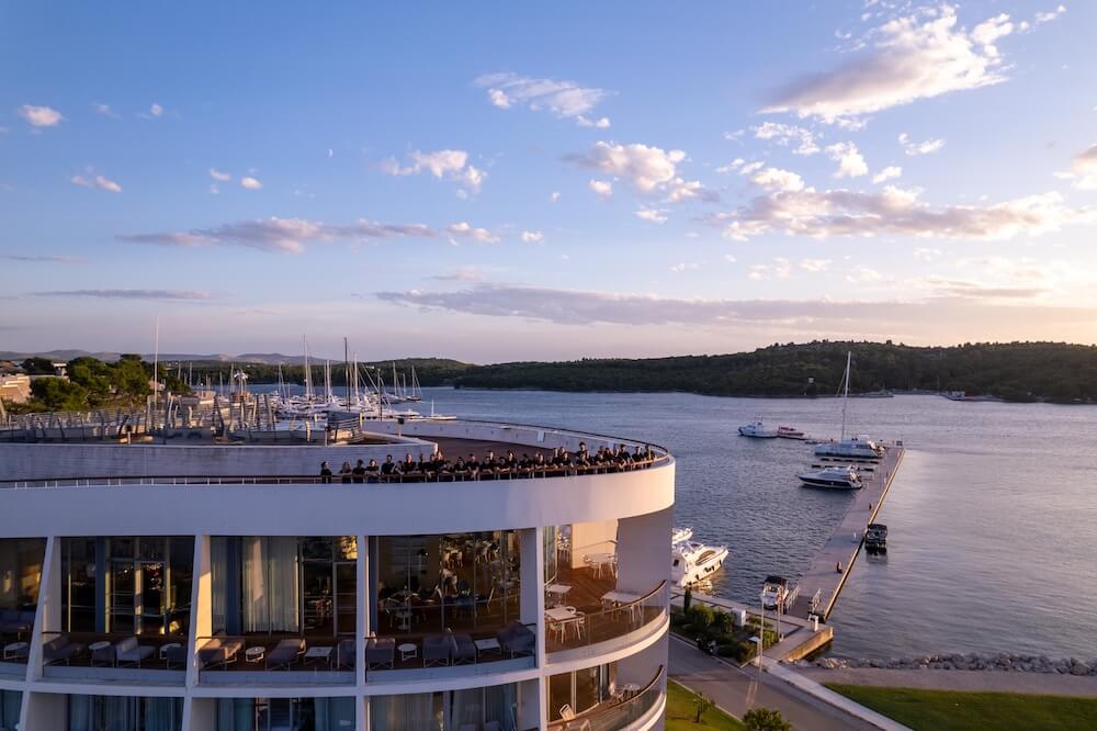 An aerial view of a modern white curved hotel building with people on the rooftop terrace, overlooking a calm bay with a pier and yachts at sunset.
