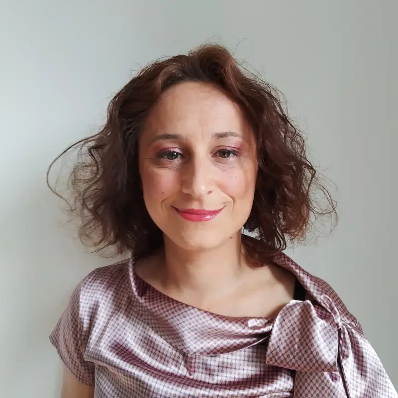 Adriana Brancaleone, a white woman with short curly hair wearing a pink blouse, pink lipstick and soft pink eyeshadow smiling in a white background.