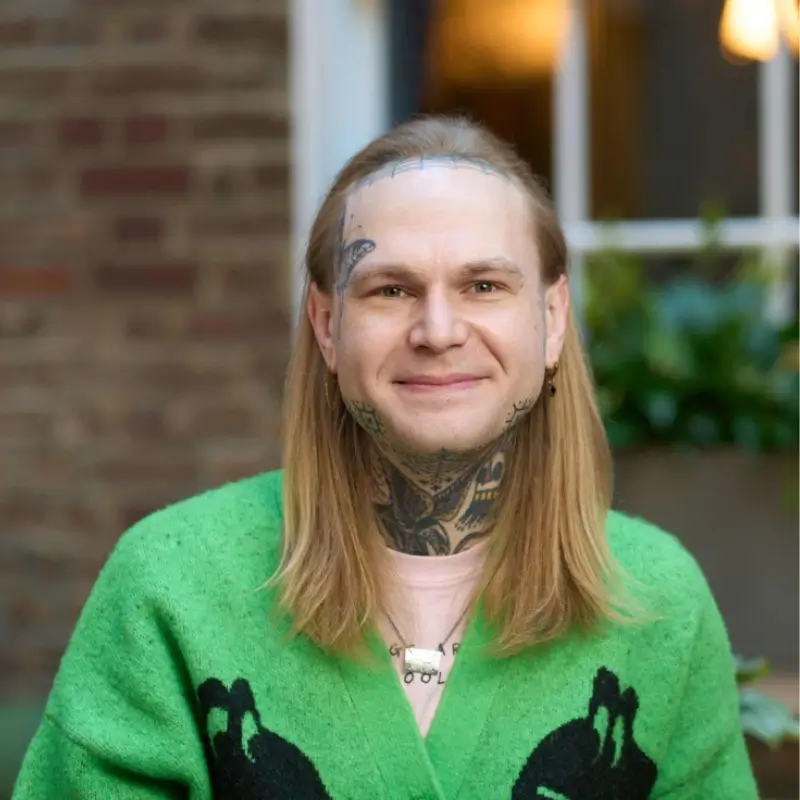 Lewis Reynolds, a white person with long blond hair wearing a neon green v neck sweater with a pink shirt under it smiling in front of a blurry brick wall. They have some neck tattoos, the most visible ones being a bird and a skull as well as a butterfly tattoo on the right side of their face. Their ears are modified to be pointy and elf-like.