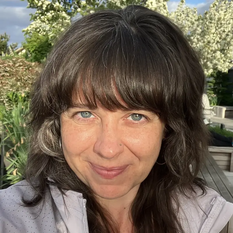 Renee Lynch, a white brunette with bangs and blue eyes, wearing a white jacket and smiling in front of greenery.