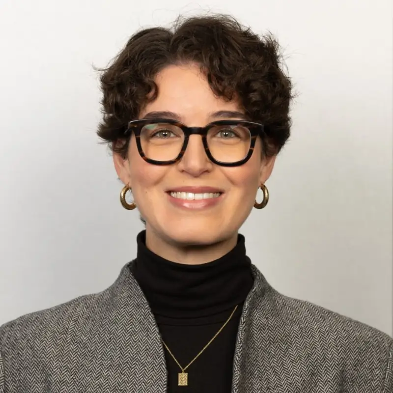 Hilal Tekmen, a brunette with short curly hair wearing square glasses, light pink lipstick, small hoop earrings and a black turtleneck with a grey coat, smiling in a white background