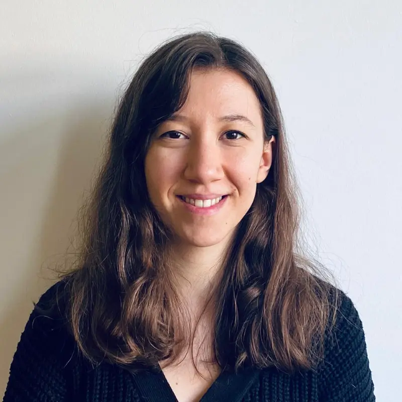 Ezgi Kan, a brunette with long wavy hair wearing a black shirt, smiling to the camera in front of a white background