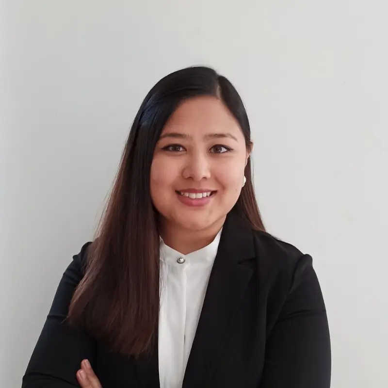 Pragati Bajracharya, an asian brunette, with long straight hair wearing a white button up shirt and a black blazer smiling with her arms crossed in front of a white background