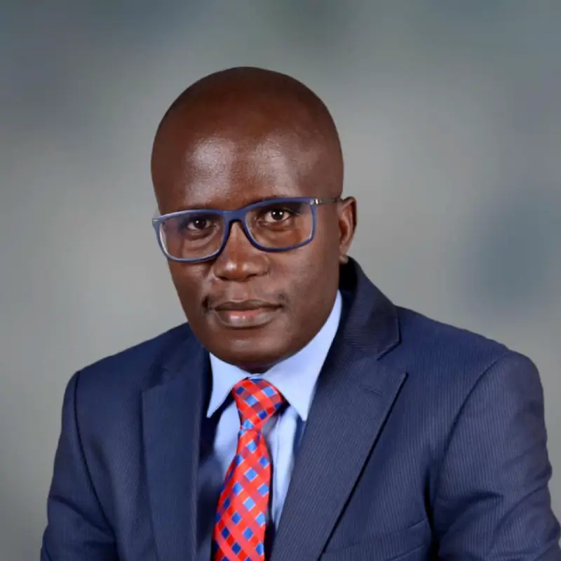 Washington Opiyo Sati, a black bald man wearing square blue glasses and a blue suit with a red tie in front of a grey background