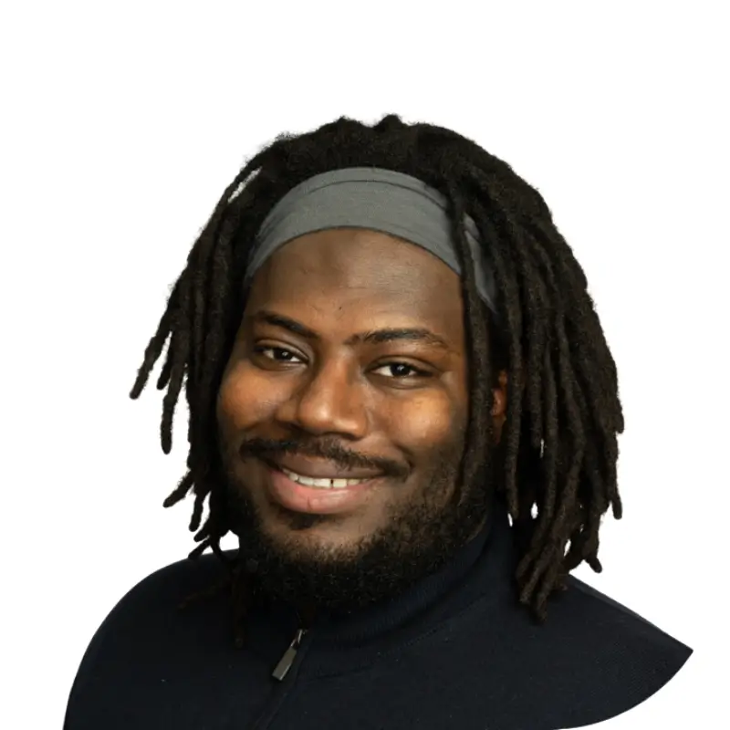 Sharif Braxton, a black man with dreads and a mustache wearing a black turtleneck and smiling in front of a white background