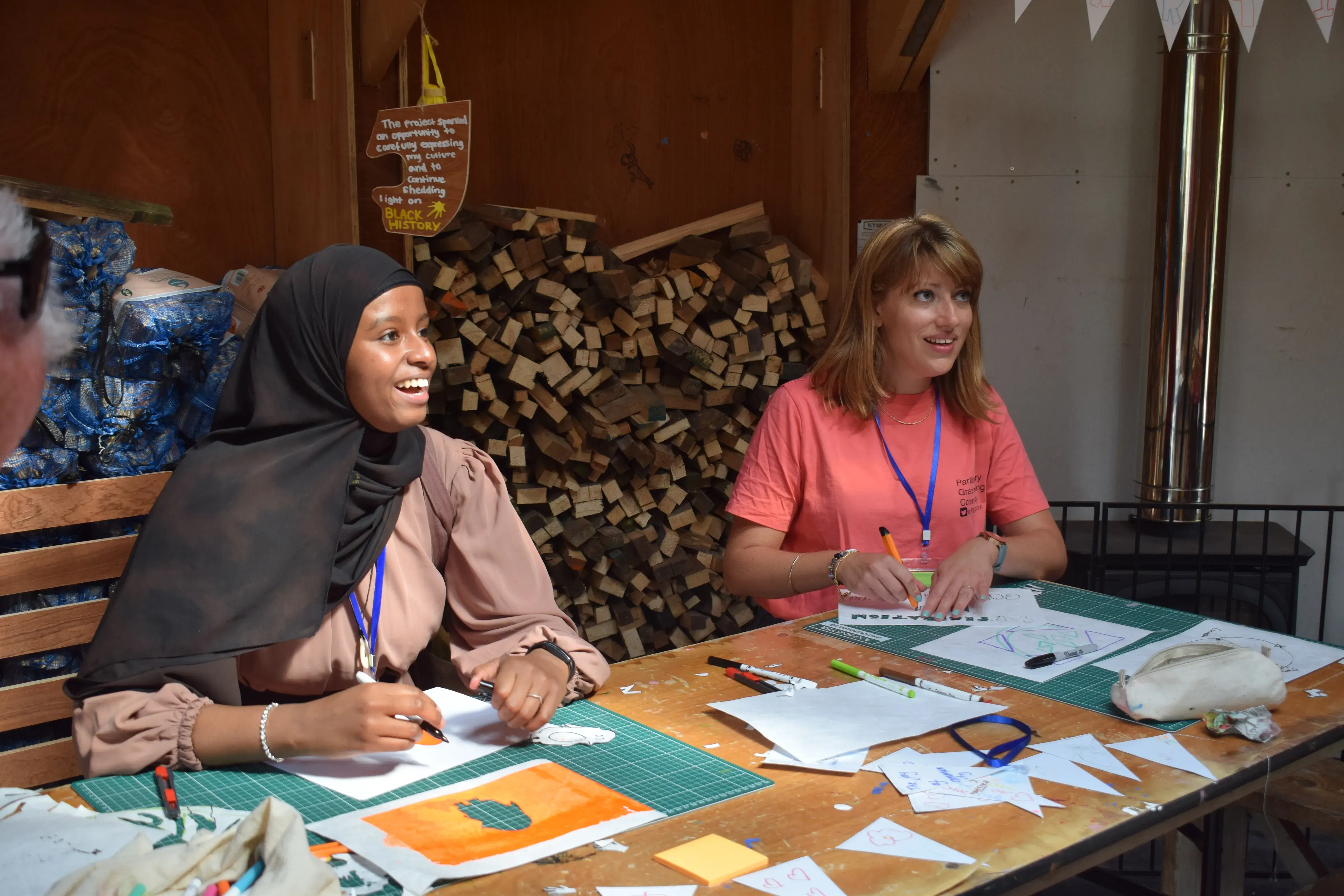 Two young women, one is wearing a black hijab and the other has light brown hair, seated at a wooden table, smiling. The table is full of art and design supplies.