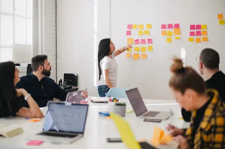 A group of people is sitting around a table with laptops in an office-like enviroment, watching a woman standing by a wall covered in colorful sticky notes.
