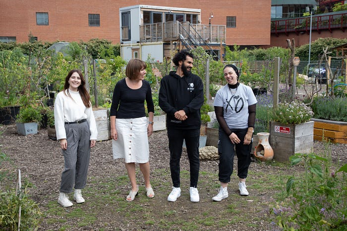 Four people stand in a community garden, smiling. From left to right: one person has long brown hair in a white blouse and gray pants, the second has short brown hair in a black top and white skirt, the third has curly black hair in a black hoodie, and the fourth has long dark hair tied back in a black shirt and pants.