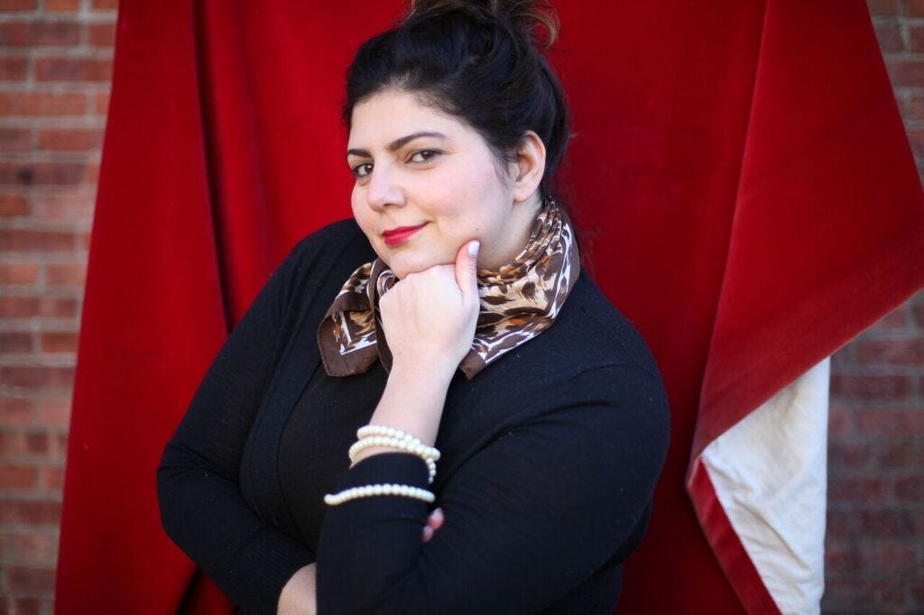 Arianne Shaffer, a white brunette, posing with a smirk and her left hand in a fist under her chin in front of a red velvet cloth hanged on a brick wall.