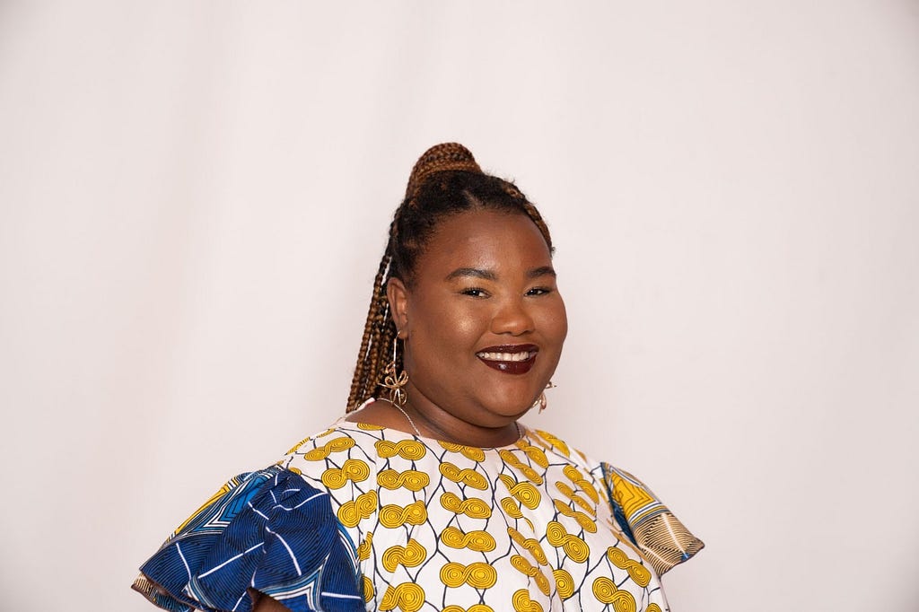 Gabrielle Bailey, a black woman wearing red lipstick with braided hair styled in a high ponytail smiling in front of a white background.