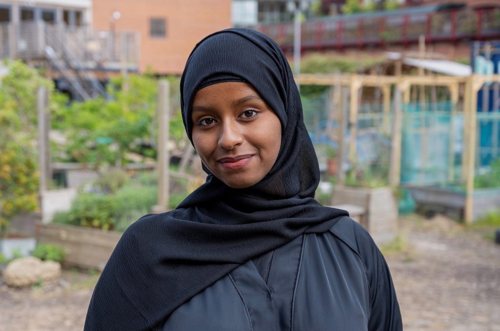 Yasmin Farah, a woman wearing a black hijab standing in front of greenery.