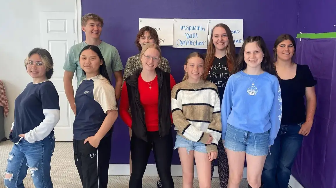 A group of nine young people stands together in a room with a purple wall. They are smiling and dressed casually, with a mix of styles. A poster in the background reads "Welcome to the Celebration."