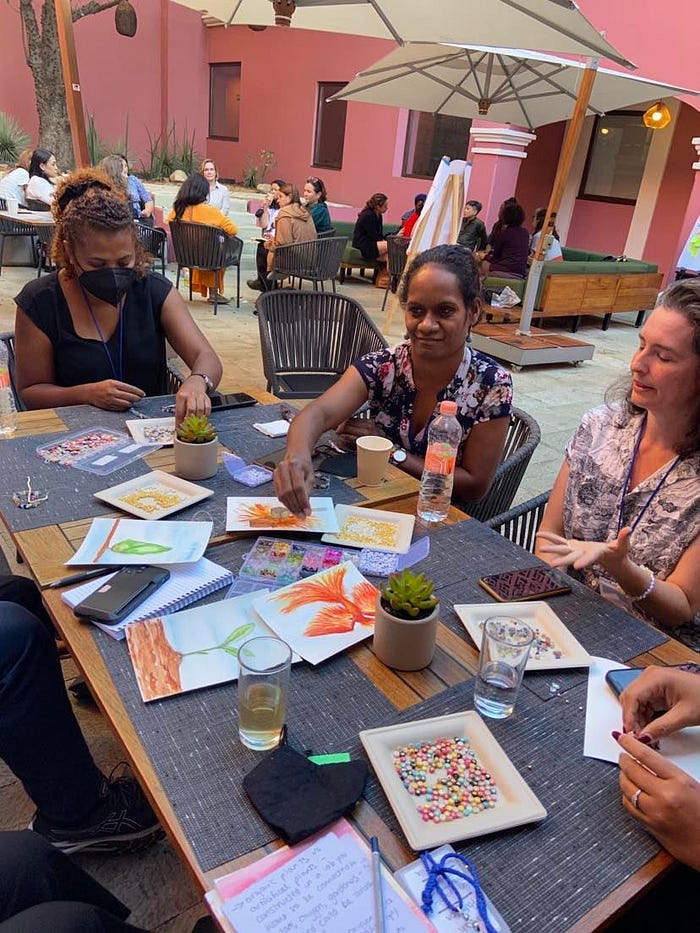 Three women participate in an arts and crafts activity at an outdoor table, with pink stone in the background.