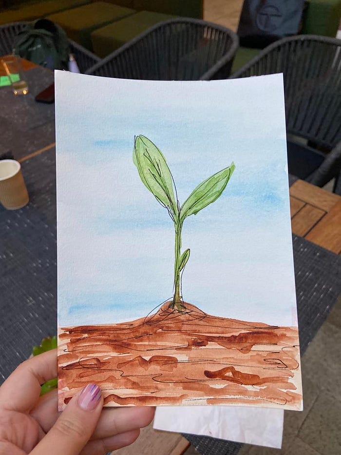A hand with pink-and-white nail polish holds a watercolor painting of a green seedling sprouting from brown earth.
