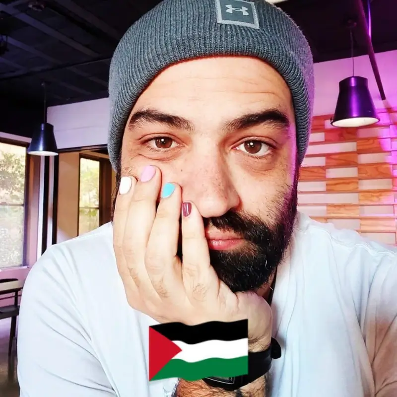 Samuel Shapiro, wearing a grey beanier and white shirt.His hands are over his beard and covering a part of his face, showing his different colored painted nails. At the bottom there is a palestinian flag emoji. 