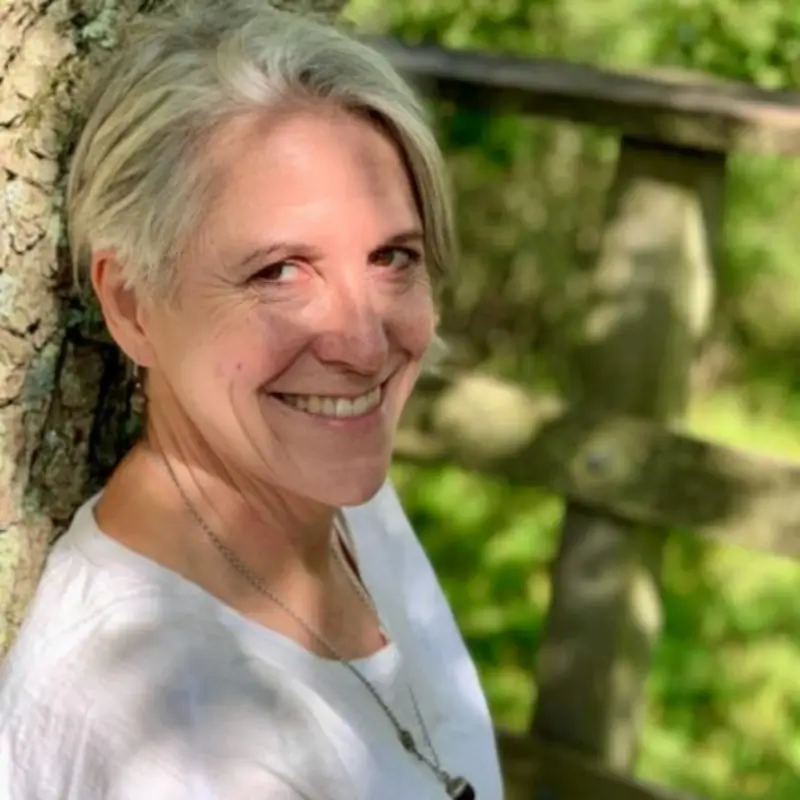 Kate Harrison, a white woman with short gray hair wearing a white top smilling while leaning against a tree, with greenery and a wooden fence in the background.